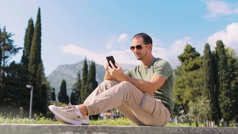 Handsome man sit and use phone smiling outdoors Stock Footage 129298643