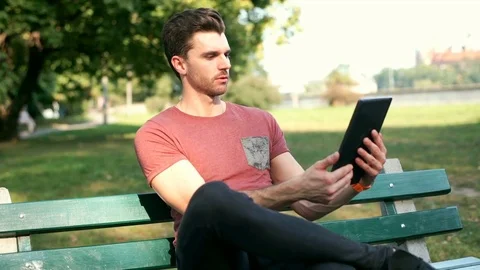 Handsome man sitting on the bench and doing selfies on tablet in the park Video stock 69964560