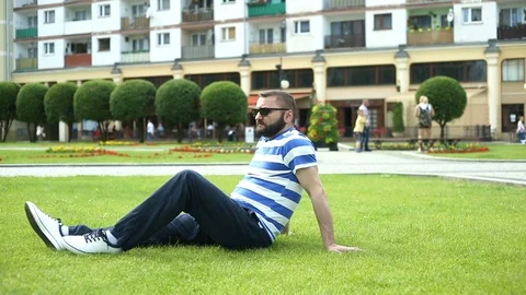 Handsome man sitting on the grass in the park and relaxing Stock Footage 76981392