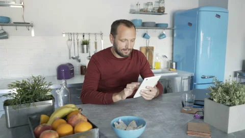 Handsome man sitting in the kitchen and browsing internet on tablet Vídeo Stock 93049177