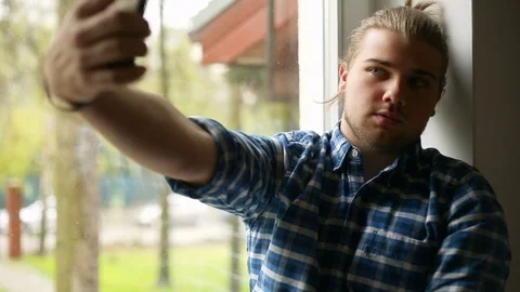 Handsome man sitting next to the window and doing selfies on smartphone Stock Footage 75136273