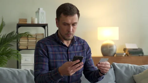 Handsome man sitting on sofa at home in living room with smartphone and credit Stock Footage 144840874