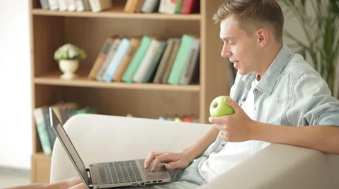 Handsome man sitting on sofa using laptop and eating apple Видео 27692009