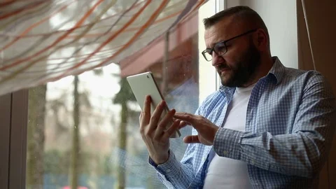 Handsome man sitting with tablet by the window at home Stock Footage 71658778