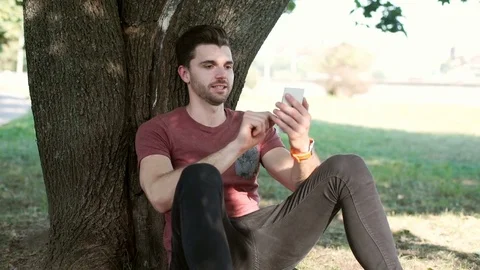 Handsome man sitting under the tree and browsing internet on smartphone Stock-Footage 69963475