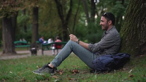 Handsome man sitting under tree, talking to friend, happily waving his hand Stock Footage 189966777