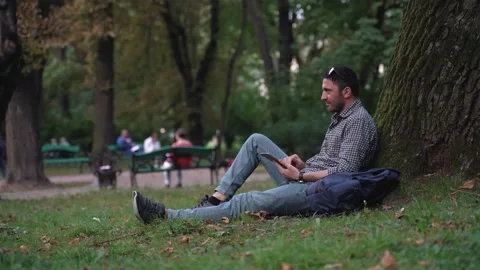 Handsome man sitting under tree, chatting with a girlfriend, wife on tablet Stock Footage 197437264