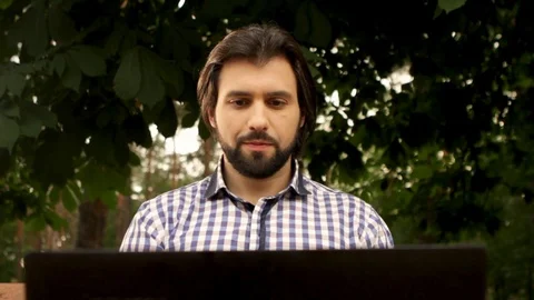 Handsome man is sitting under trees and holding laptop. He is looking at screen Stock Footage 99739966
