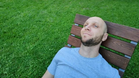 Handsome man is smiling while lying on wooden sunbed in city park Stockbeeldmateriaal 218977152