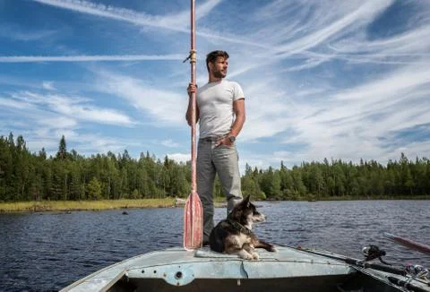 Handsome man is standing on the boat with baddle in his hands and dog near of Stock Photos