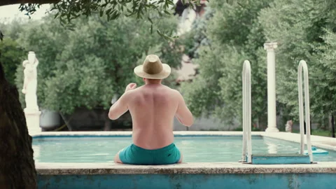 Handsome man in straw hat sitting on pool side and drinking refreshing beverage Stock Footage 261206293