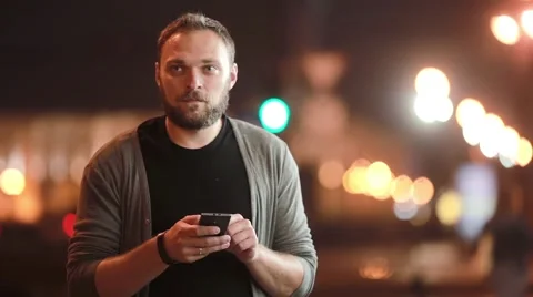 Handsome man texts, uses his smartphone and goes away. Blurred city lights in Video stock 67951931