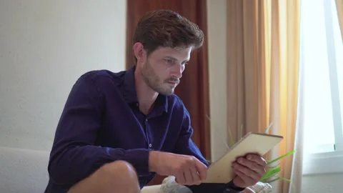 Handsome man is using a digital tablet and smiling while resting on couch at 스톡 동영상 140599006