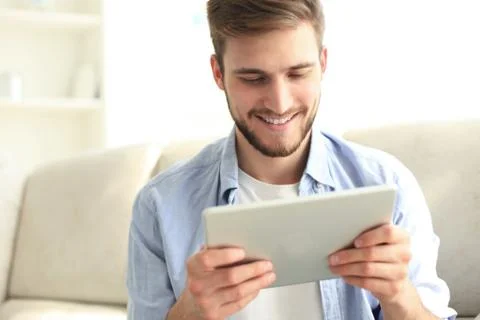 Handsome man is using a digital tablet and smiling while resting on couch. Stock Photos