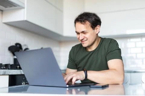 Handsome man using a laptop in the kitchen. Working from home in quarantine l Stock Photos