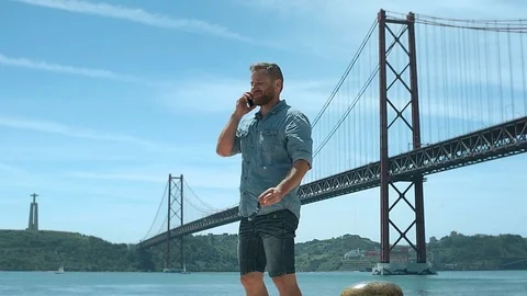 Handsome man walking next to the river while chatting on cellphone, steadycam sh Stock Footage