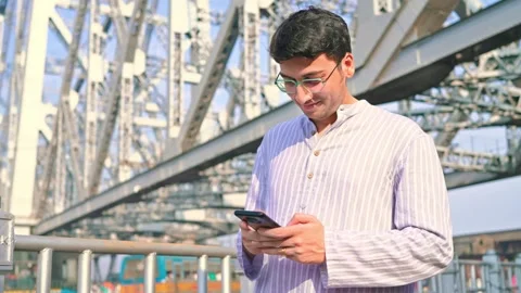 A handsome man walking under the Howrah bridge, Kolkata using a smartphone Stock Footage 268974352