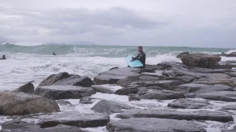 Handsome man in wetsuit with surfboard sitting on rocks in wave Stock Footage 303768008