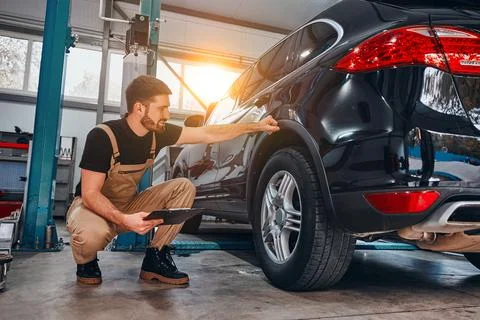 Handsome mechanic in uniform is making notes examining car in auto service. Stock Photos