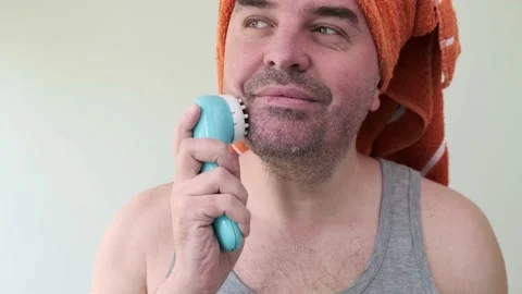 Handsome middle aged bearded man with towel on head using portable face massager Stock Footage 130564281