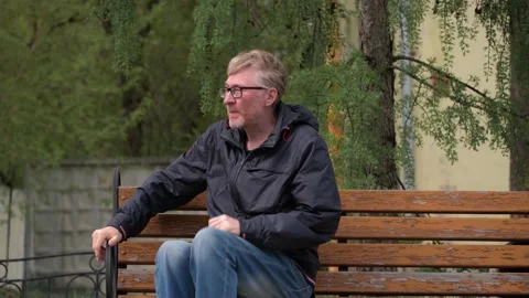 Handsome middle aged man in blue jacket and jeans approaches bench and sits down Stock Footage 250497610