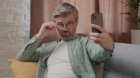 Handsome middle aged man with grey hair sitting on the sofa using phone taking Vídeos de archivo 220201472