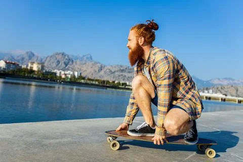Handsome redhaired man with long beard skateboarding near river at sunny summer Stock Photos