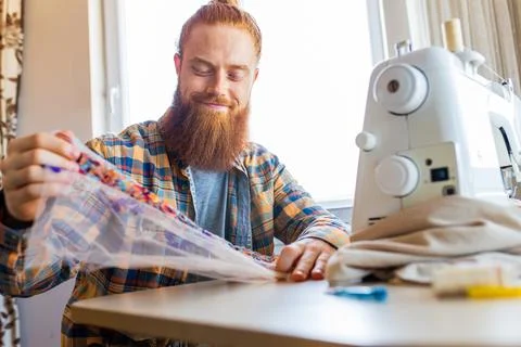 Handsome redhaired man with long beard sews at a sewing machine at home studio Stock Photos
