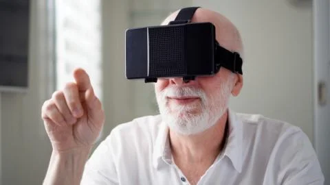 Handsome senior man in white using VR 360 glasses at home. Making browse and Stock Photos
