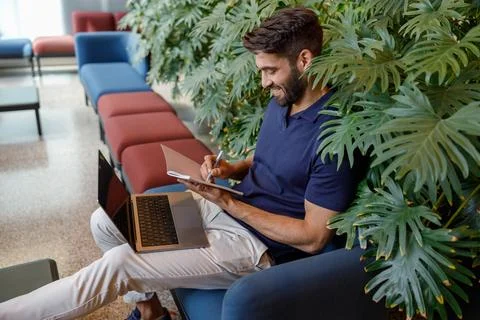 Handsome smiling freelancer is making notes during work on laptop in office hall Stock Photos