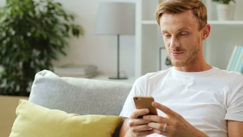 Handsome smiling man in white t-shirt sitting on the sofa and using smartphone Stock Footage 79997813