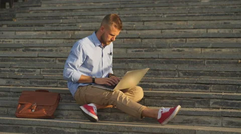 Handsome student sitting on stone steps and using a laptop Stock Footage 68067871