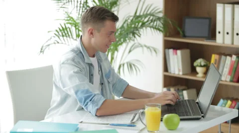 Handsome student sitting at table writing in notebook using laptop and drinking Stock Footage 27692049