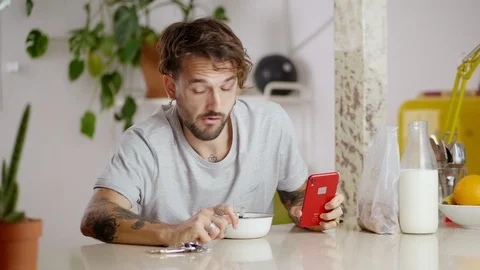 Handsome tattooed guy eating corn flakes breakfast using smartphone and runs Stock Footage 108795826