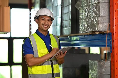 Handsome warehouse worker wearing security vest using digital tablet while Stock Photos