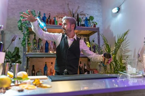 Handsome young bartender inside the bar skillfully throws a shaker with ice and Stock Photos