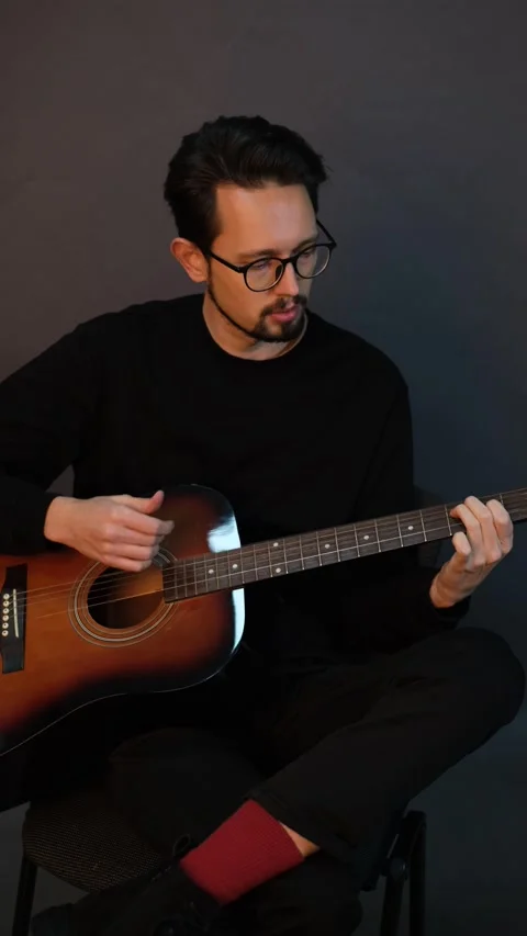 A handsome young brunet man playing guitar while sitting on a chair in a dark st Stock Footage 320356090