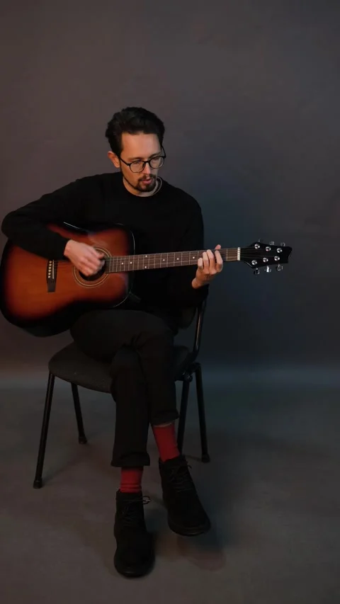 A handsome young brunet man playing guitar while sitting on a chair in a dark st Stock Footage 320356091