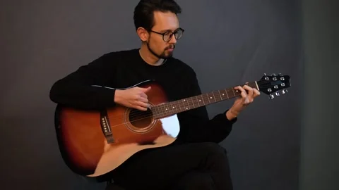 A handsome young brunet man playing guitar while sitting on a chair in a dark st Stock Footage 320356196