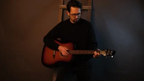 A handsome young brunet man playing guitar while sitting on a chair in a dark st Stock Footage 320356805