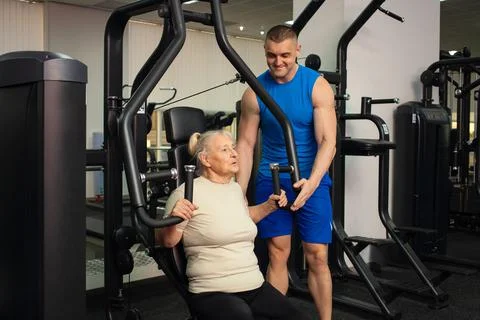 A handsome young coach man helps an old woman train sports in gym. Happy peop Stock Photos