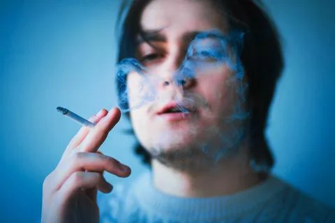A handsome young dark-haired guy with a white sweater smokes a cigarette, exh Stock Photos