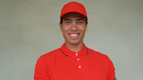 Handsome young delivery worker in red uniform is holding a pizza, looking at Stock Footage 112831045