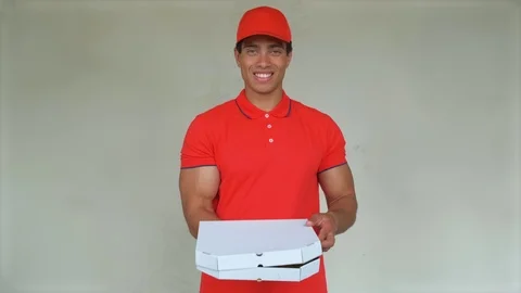 Handsome young delivery worker in red uniform is holding a pizza, looking at Stock Footage 112831077