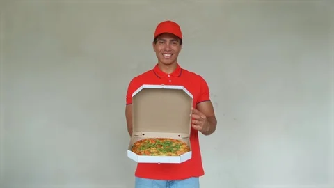Handsome young delivery worker in red uniform is holding a pizza, looking at Stock Footage 112831118
