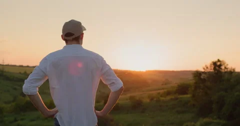 Handsome young farmer stands in front of a picturesque countryside as the sun Stock Footage 252015298