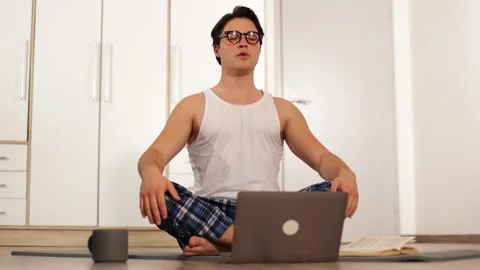Handsome young guy doing yoga on a mat on the floor at home front of computer 動画素材 253827085