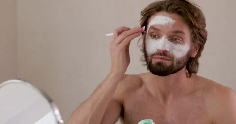 Handsome young guy looking at small mirror and applying facial mask with brush Stock Footage 142529838