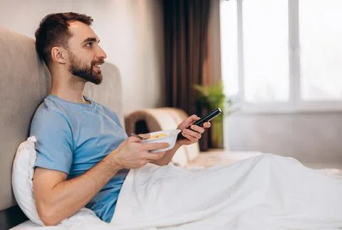 Handsome young man with beard having breakfast in bed and watching tv. weekend Stock Photos
