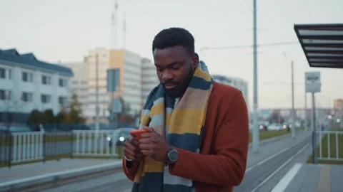 Handsome young man in coat using smartphone tracking tram online waiting at Stock Footage 121476412